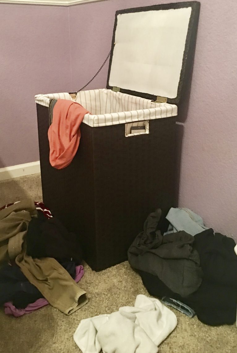 A hamper is surrounded at is base with clothes. One garment hangs out of the side of the hamper.